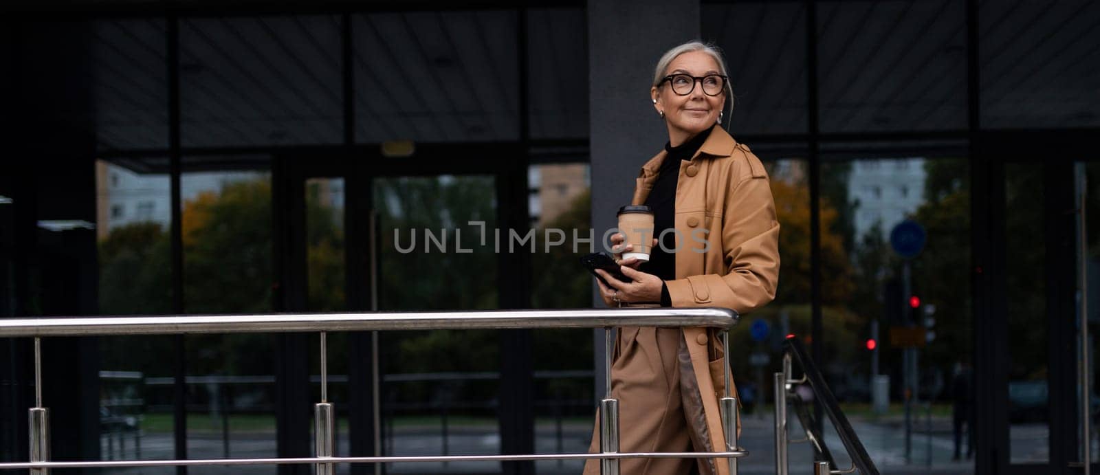 horizontal photo of a successful mature adult woman boss with a mug of coffee in her hands on the background of the building by TRMK