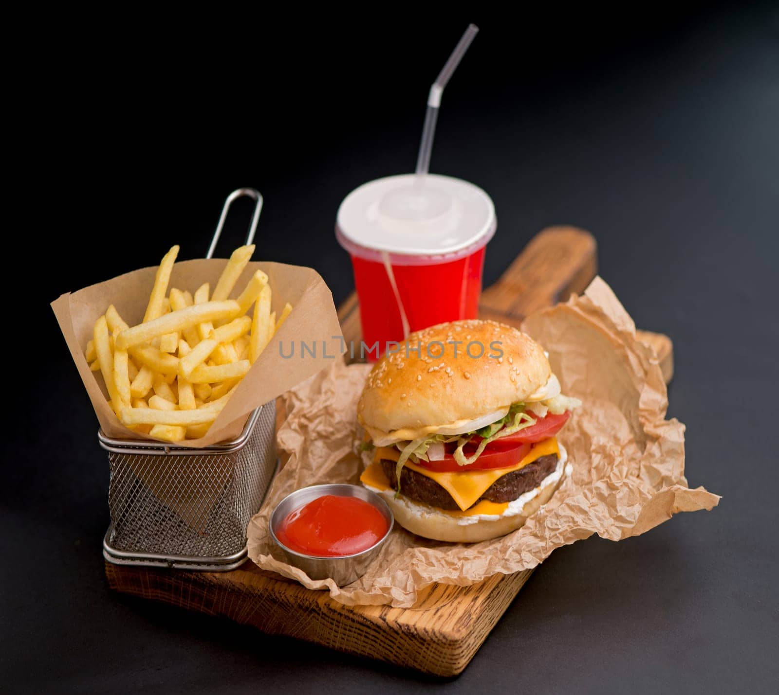 Classic burgers with becon, meat, cheese, onion, tomato and cheese and integral bun. On textured black board. Close up. Fried potatoes and glass of cola.