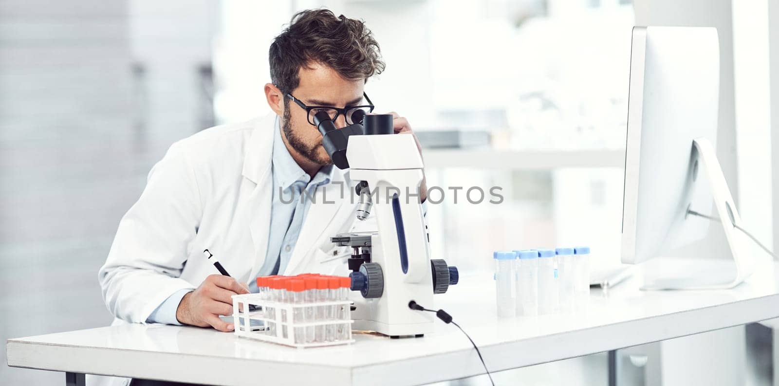 Doing what medical professionals do best. a focused young male scientist making notes while looking trough a microscope and being seated inside a laboratory. by YuriArcurs