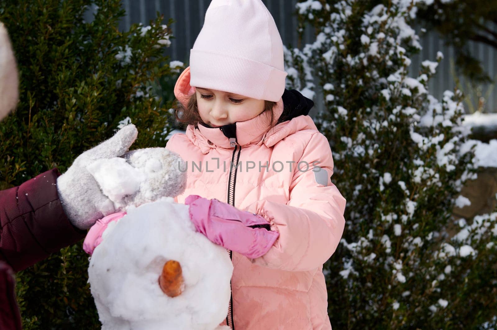 Cute little child girl in warm pink down jacket, enjoying making a snowman in a backyard, spending happy winter holidays by artgf