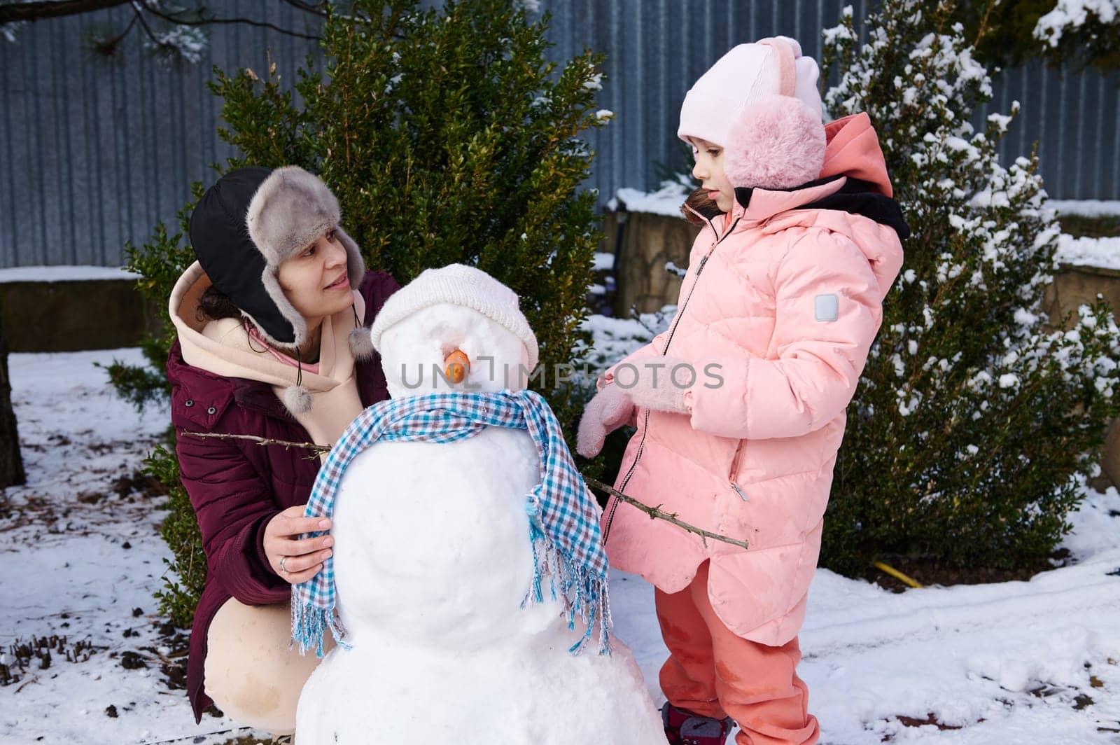 Happy loving mother and adorable her daughter spend wonderful time together, building snowman in snow covered backyard. by artgf