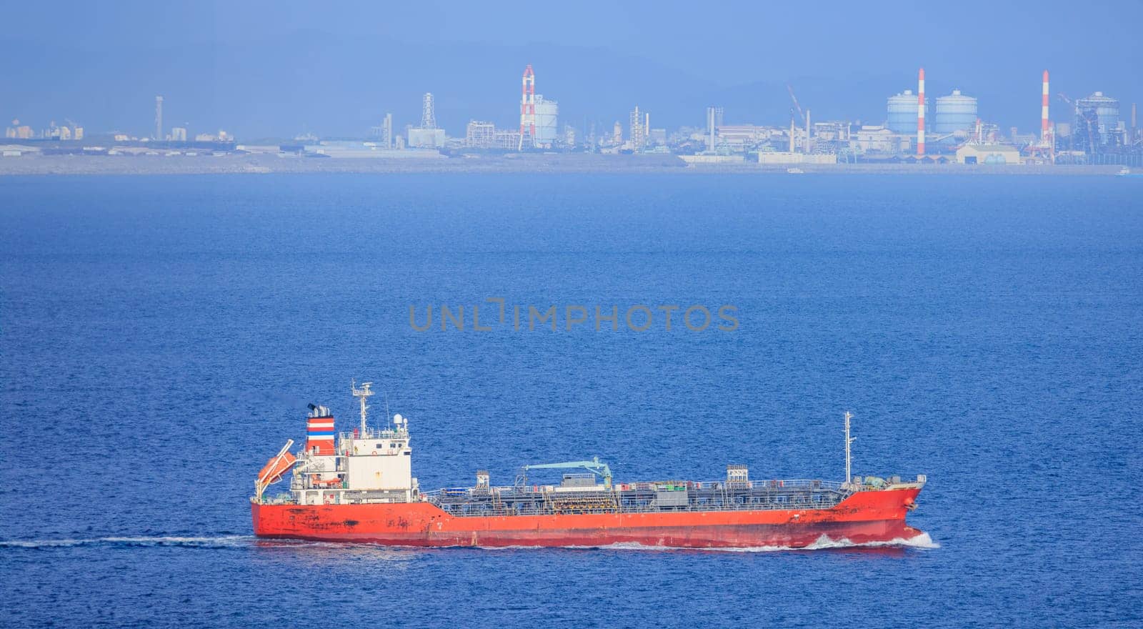 Small red cargo ship sails past chimneys at industrial refinery on coast. High quality photo