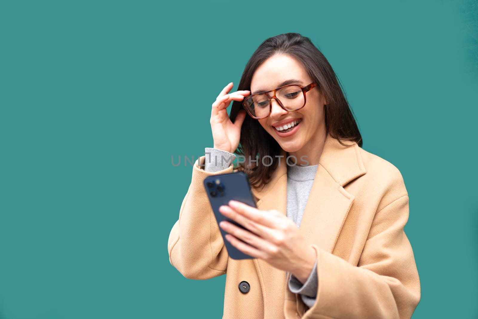 Excited woman in glasses holding smartphone in hand dressed stylish trench coat standing isolated on green background