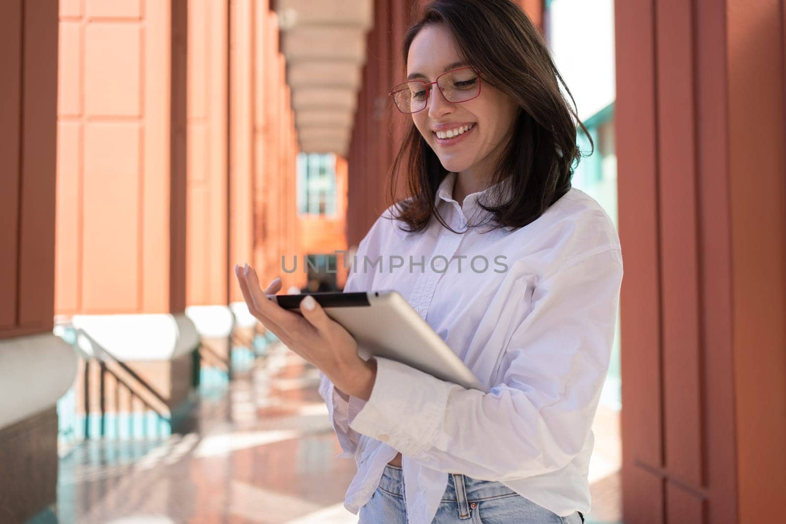 Businesswoman in glasses using digital tablet touching screen and smile, standing outdoor dressed white shirt. Female freelancer holding tablet while walk outside