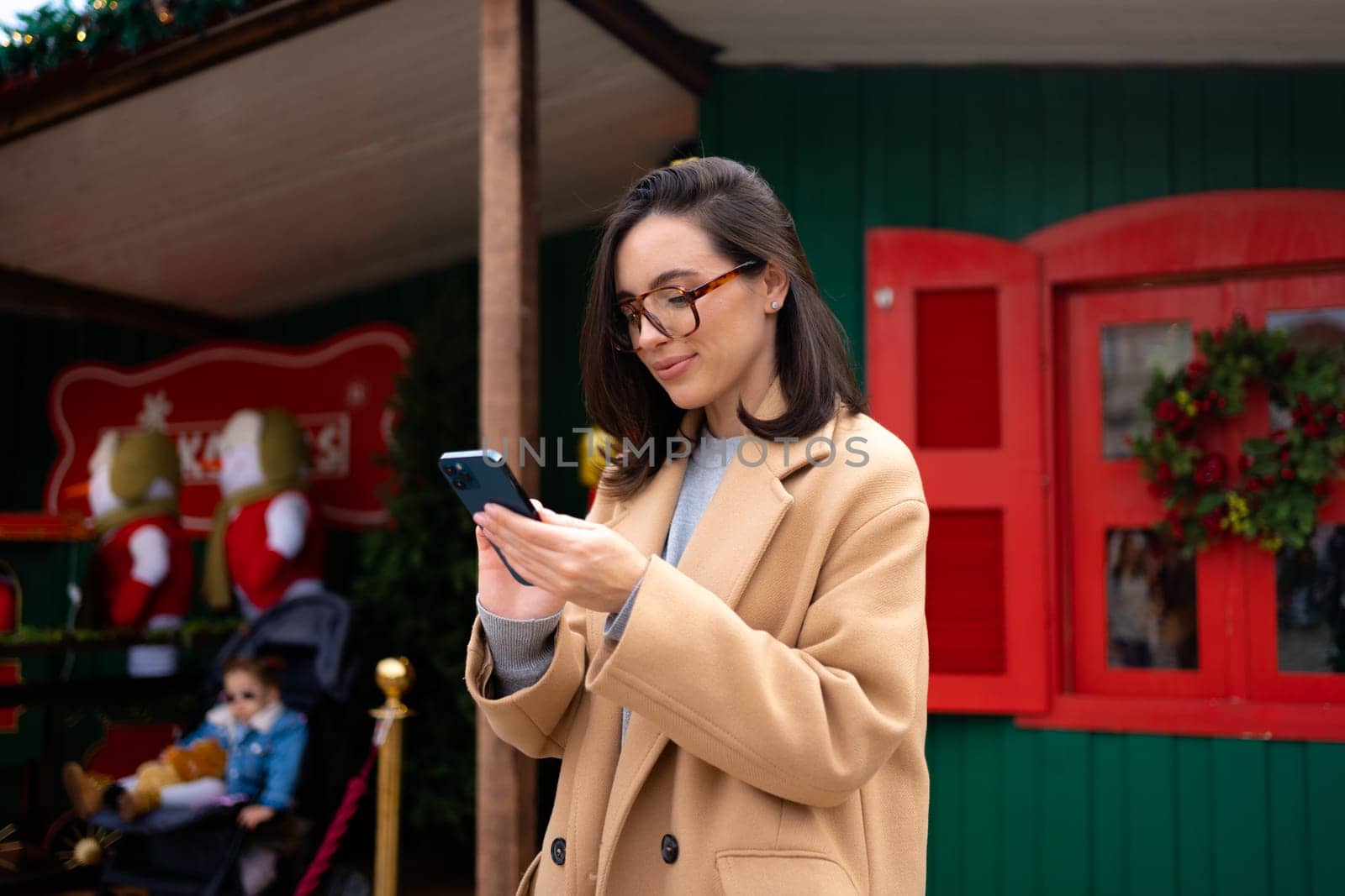 Smiling business woman wearing trench coat standing on Christmas city street using smartphone applications on cell phone, reading news on smartphone, fast connection, checking mobile apps outdoors.