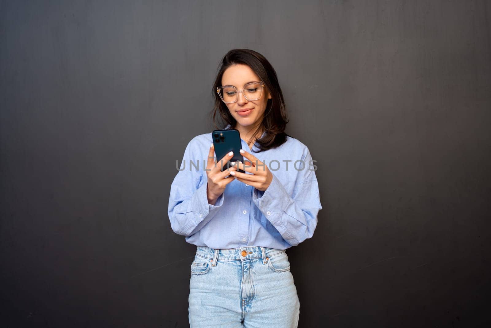 Satisfied hipster girl wear glasses, types text message on mobile phone, enjoys online communication, types feedback, wears denim shirt, isolated on black studio background. Technology concept