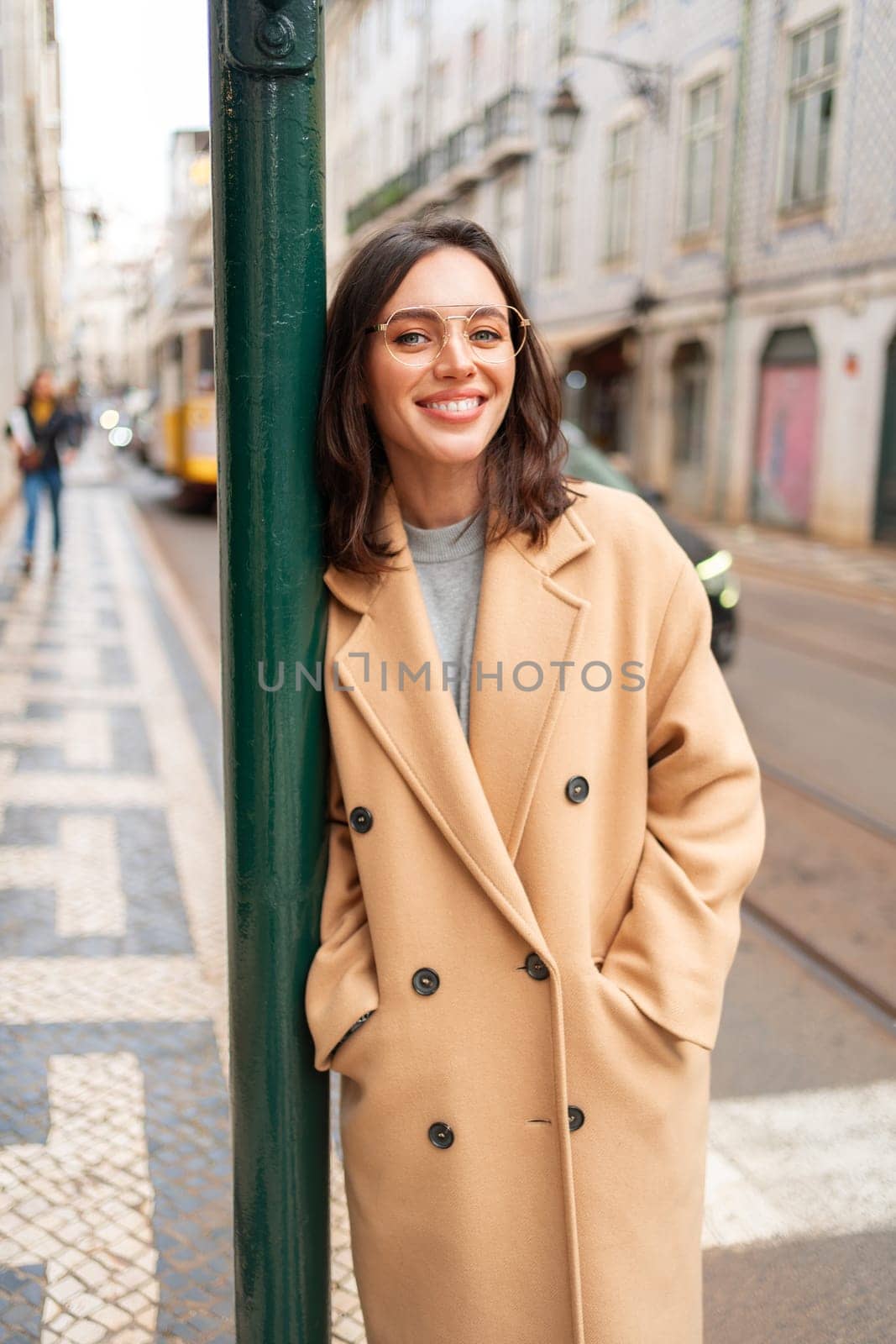 Happy caucasian girl walking city street on sunny autumn day. Brunette woman wears stylish trench coat and glasses. Positive emotions lifestyle concept