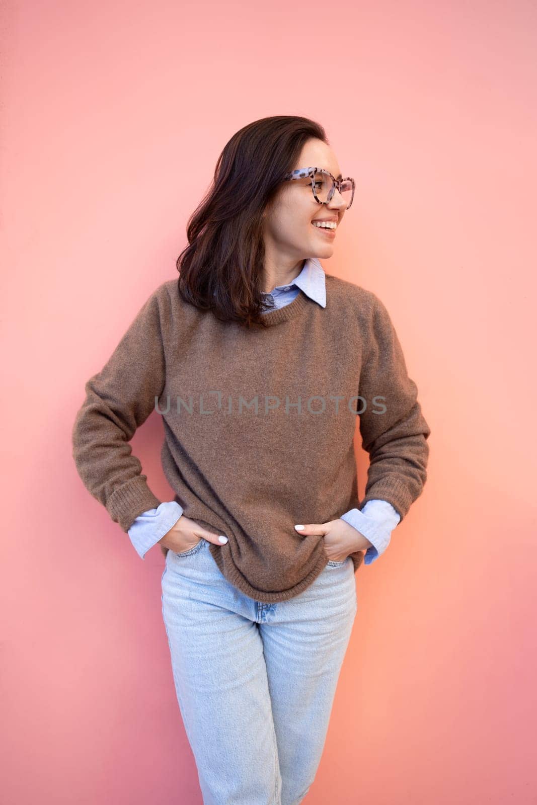 Happy woman in glasses outdoor on yellow and pink color background. Positive people concept. Smiling girl looking side, hands in pocket, dressed sweater and jeans. Vertical photo