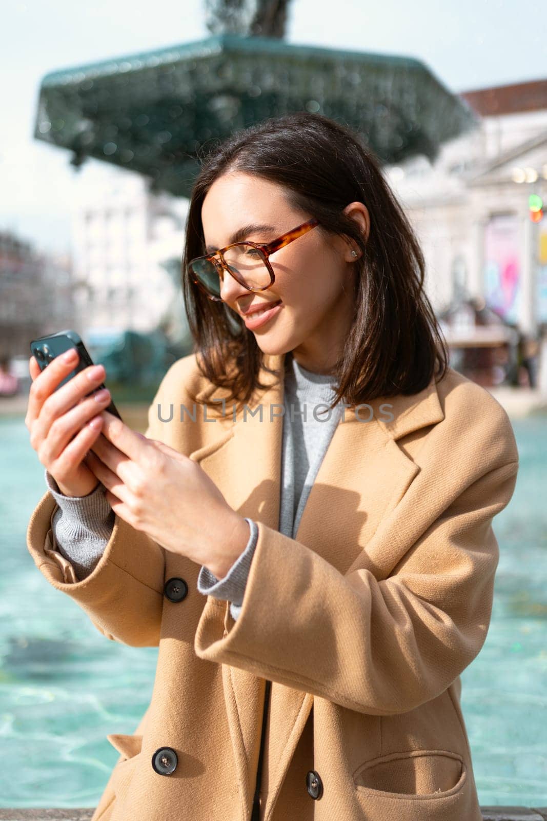 Woman wearing glasses, sitting at the fountain, holding smartphone and smiling sweetly. Good vibes and feelings. Online communication using mobile digital device. Portugal, Lisbon