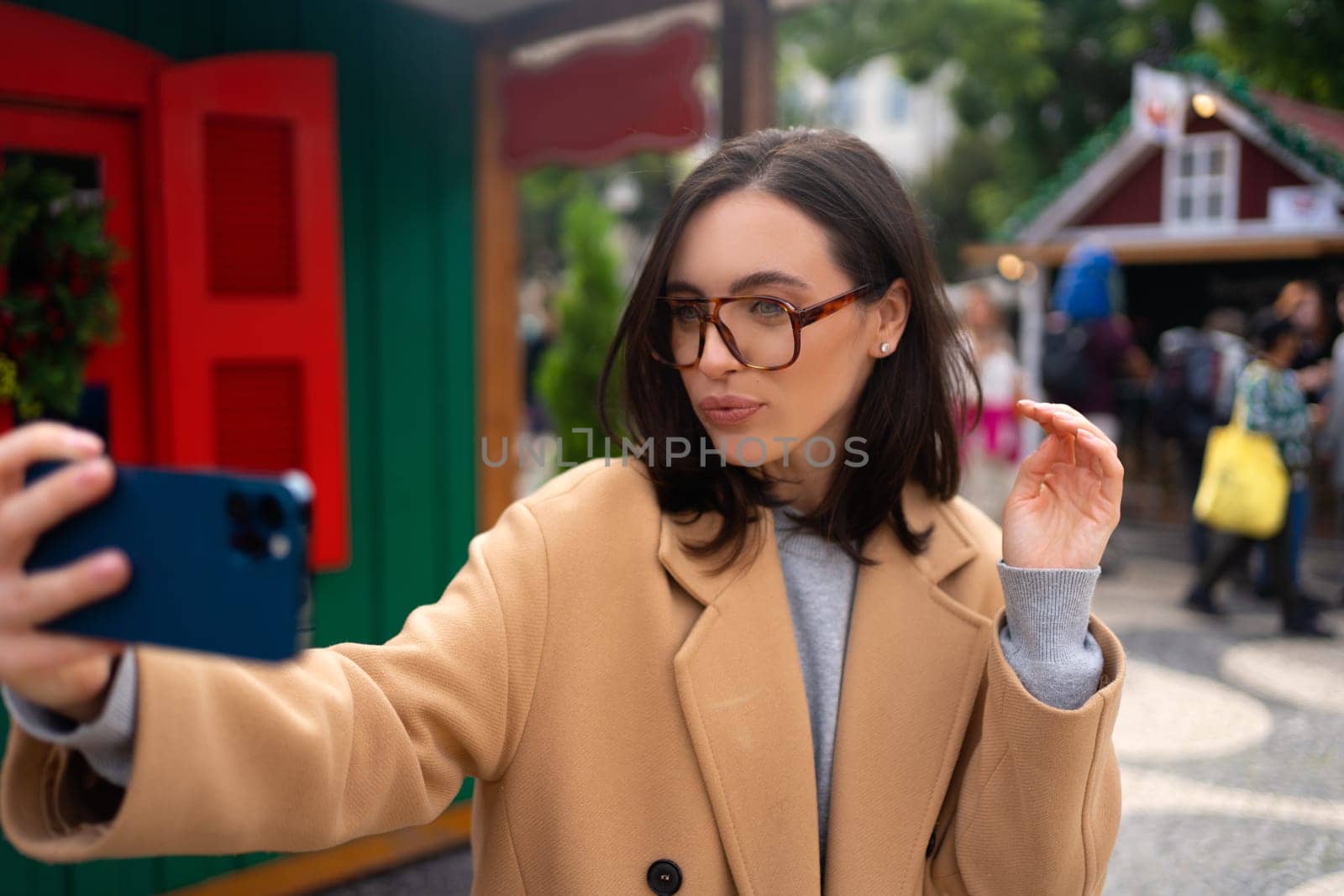 Portrait of positive woman 30s in glasses laughing while taking selfie photo on smartphone dressed stylish trench coat standing outside on autumn city street.