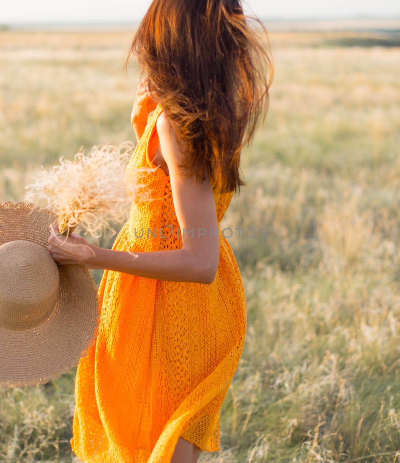 Beauty romantic girl outdoors. Rear view of a beautiful girl dressed in a casual orange dress with a straw hat in her hands on a field in the sunlight. Blows long hair. Autumn. Shine the sun, sunshine. backlit Warm tinted by Ekaterina34