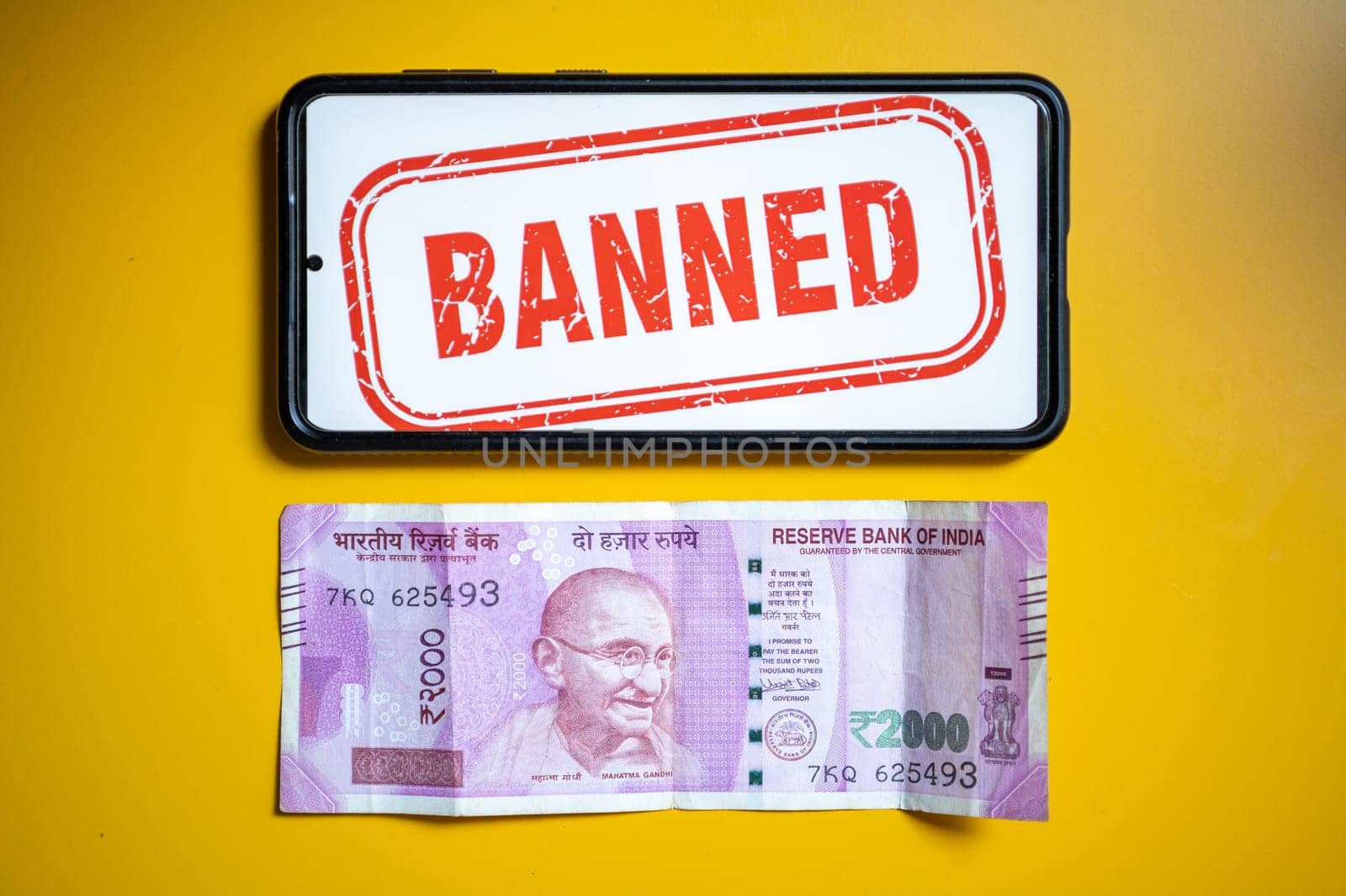 Rs 2000 Indian bank note that has been removed from circulation through demonitization on yellow table with mobile showing banned callout in India