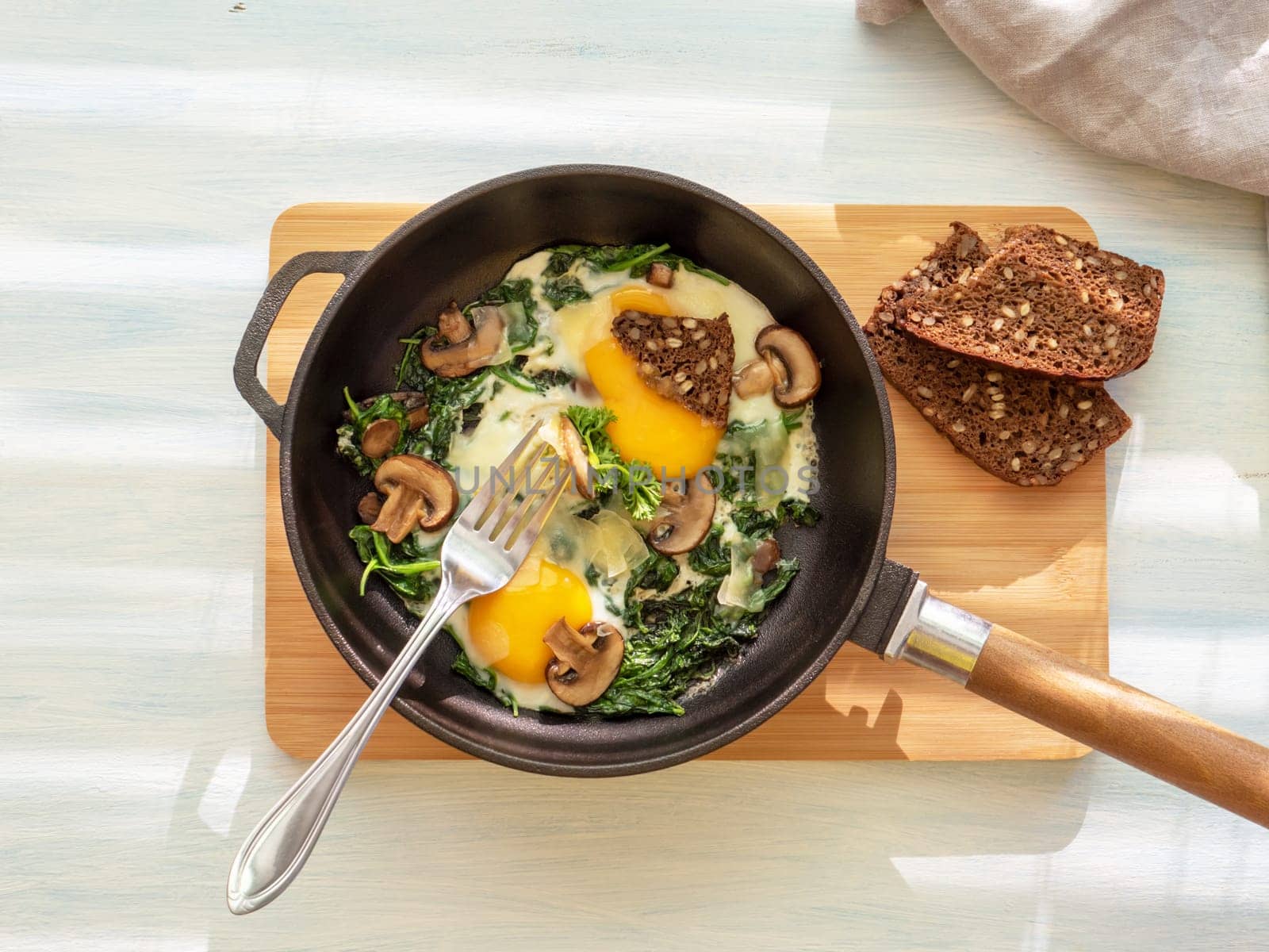 Fried eggs with spinach, mushrooms, cheese and bread in cooking pan . Healthy homemade dish for low carb diet on a light blue wooden table with linen towel. flat lay. Top view. Idea for breakfast.