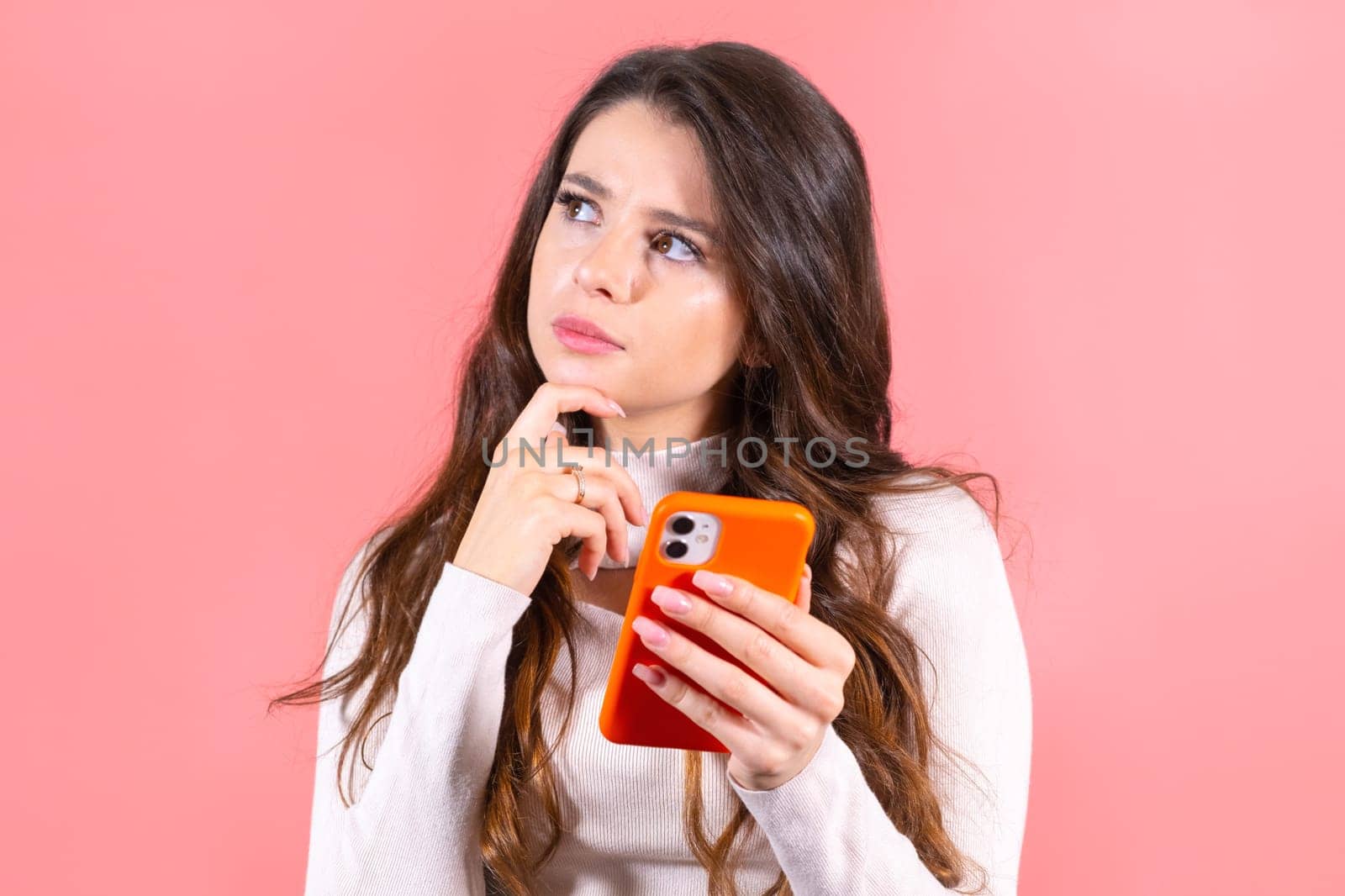 Young woman with amused and smiling expression enjoys scrolling social media on orange smartphone. Long-haired brunette lady smiles looking at feeds