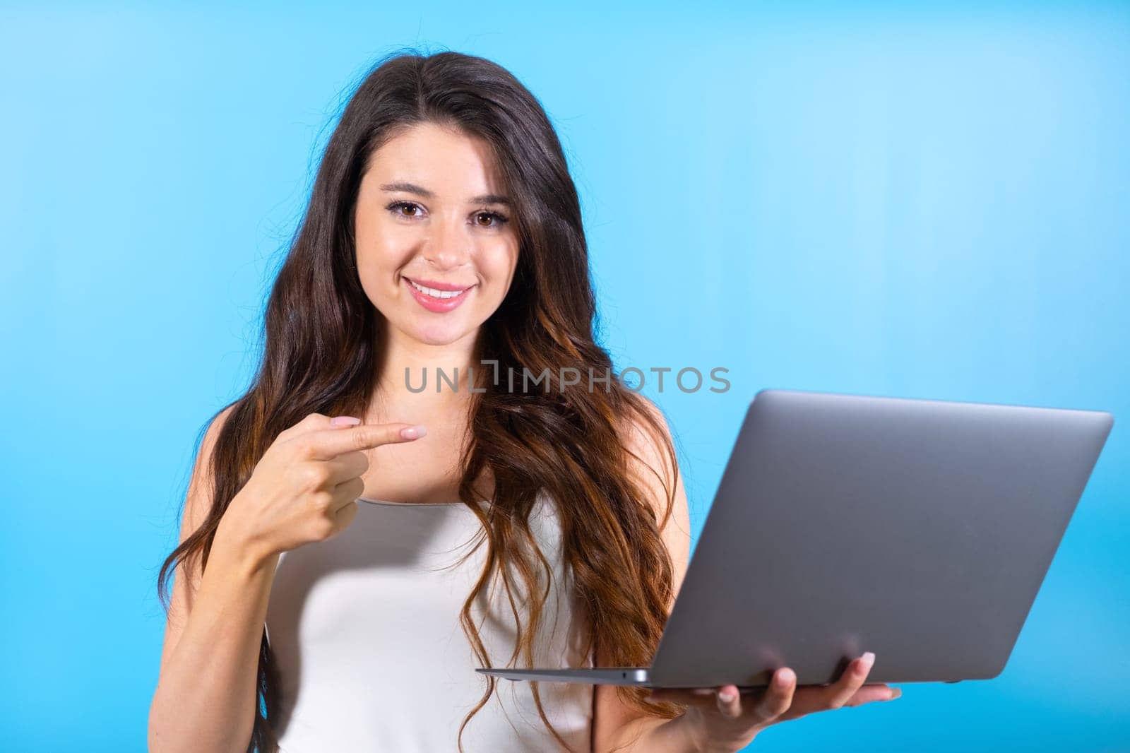 Brunette woman with amused and delighted expression looks at screen of laptop. Lady points to modern compact laptop with money win in blue studio