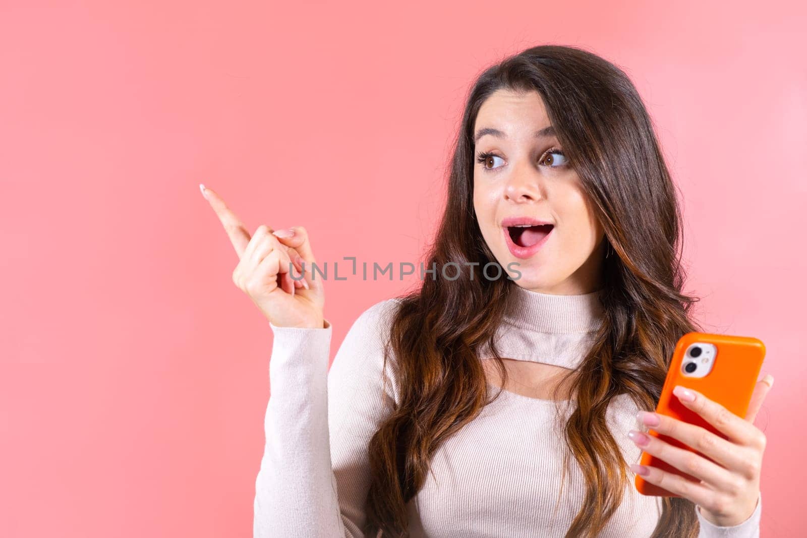 Young woman with delighted expression looks at smartphone pointing finger to wall. Brown-haired lady with amused expression at pink studio wall