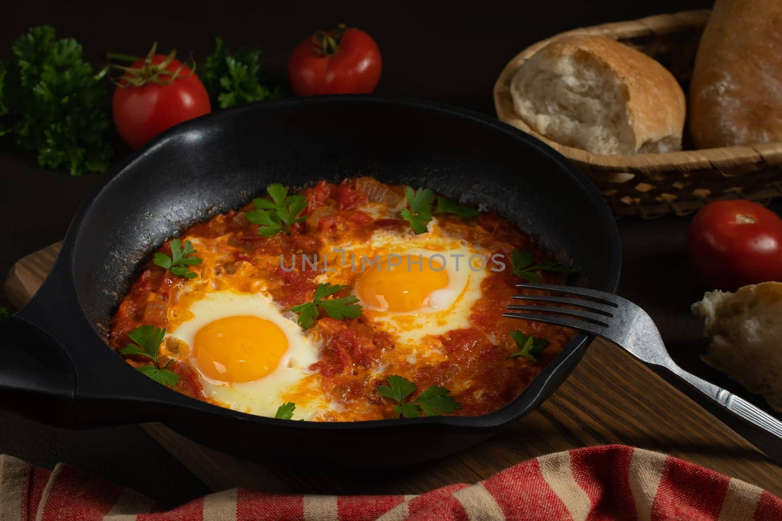 Shakshuka from two eggs in tomato sauce with fresh tomatoes, spices and herbs in a black frying pan. Close-up scrambled eggs.