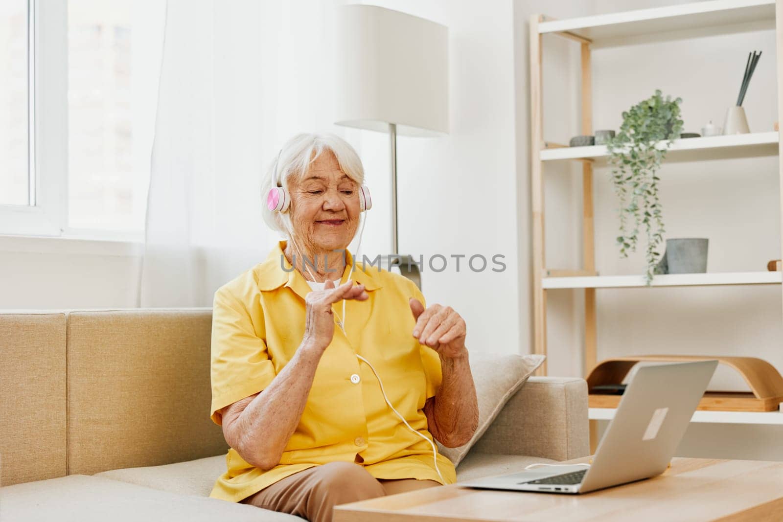 An elderly woman wearing headphones with a laptop communicating online by video call, sitting on the couch at home and working in a yellow shirt in front of a window, the lifestyle of a retired woman. High quality photo