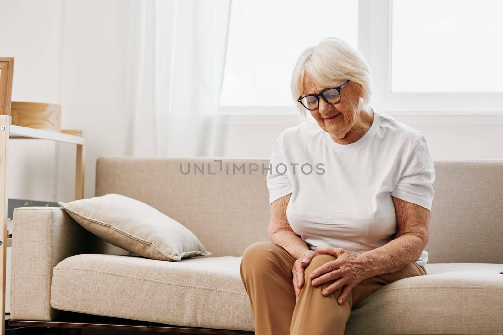 Elderly woman severe pain in her leg sitting on the couch, health problems in old age, poor quality of life. Grandmother with gray hair holds on to her sore knee, problems with joints and ligaments. by SHOTPRIME