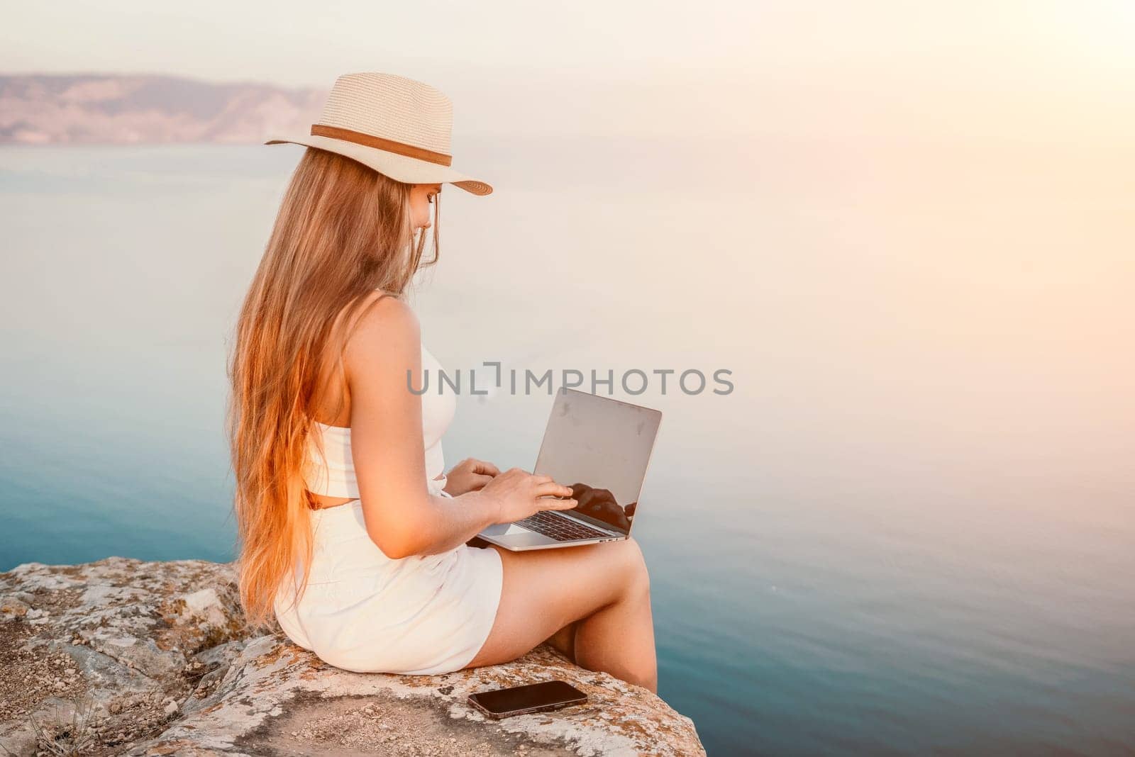 Digital nomad, Business woman working on laptop by the sea. Pretty lady typing on computer by the sea at sunset, makes a business transaction online from a distance. Freelance, remote work on vacation