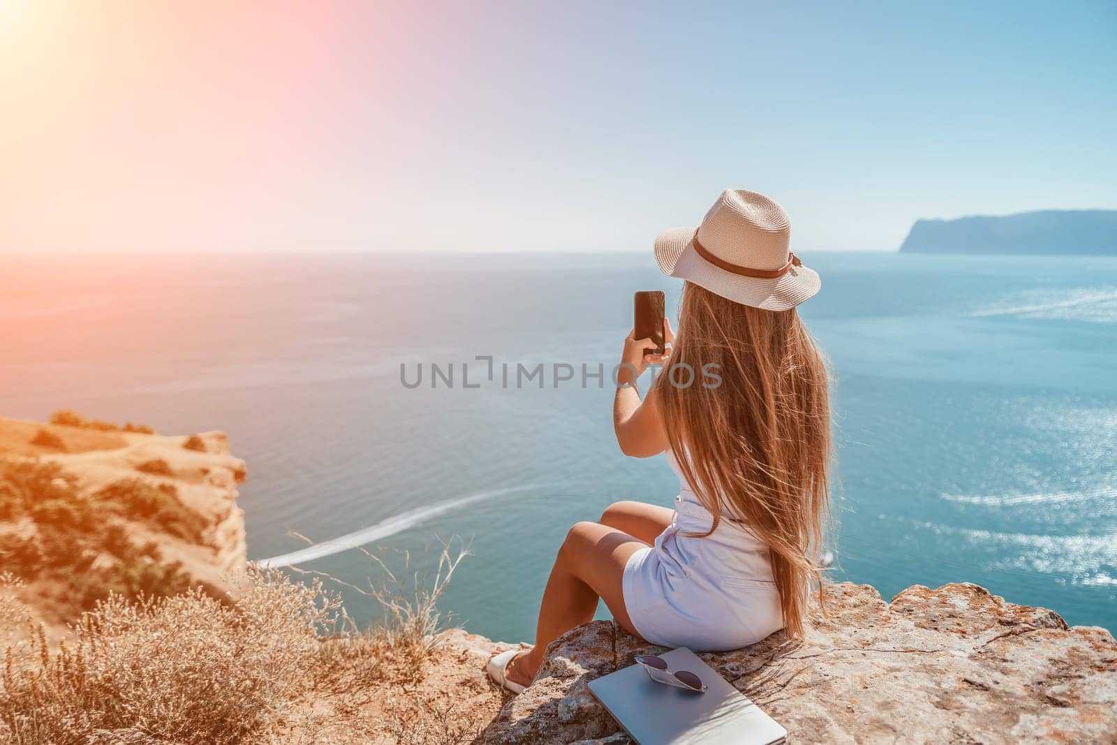 Successful business woman in yellow hat working on laptop by the sea. Pretty lady typing on computer at summer day outdoors. Freelance, travel and holidays concept.