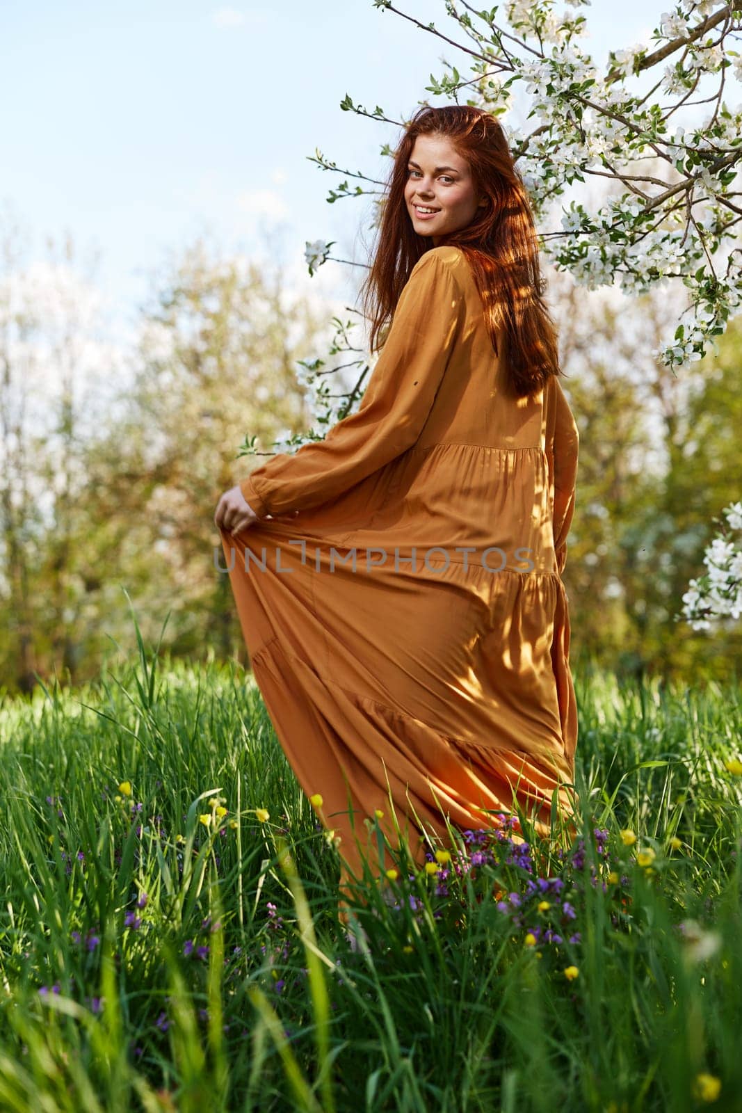 a sweet, modest, attractive woman with long red hair stands in the countryside near a flowering tree and lifts the hem of her dress up by Vichizh