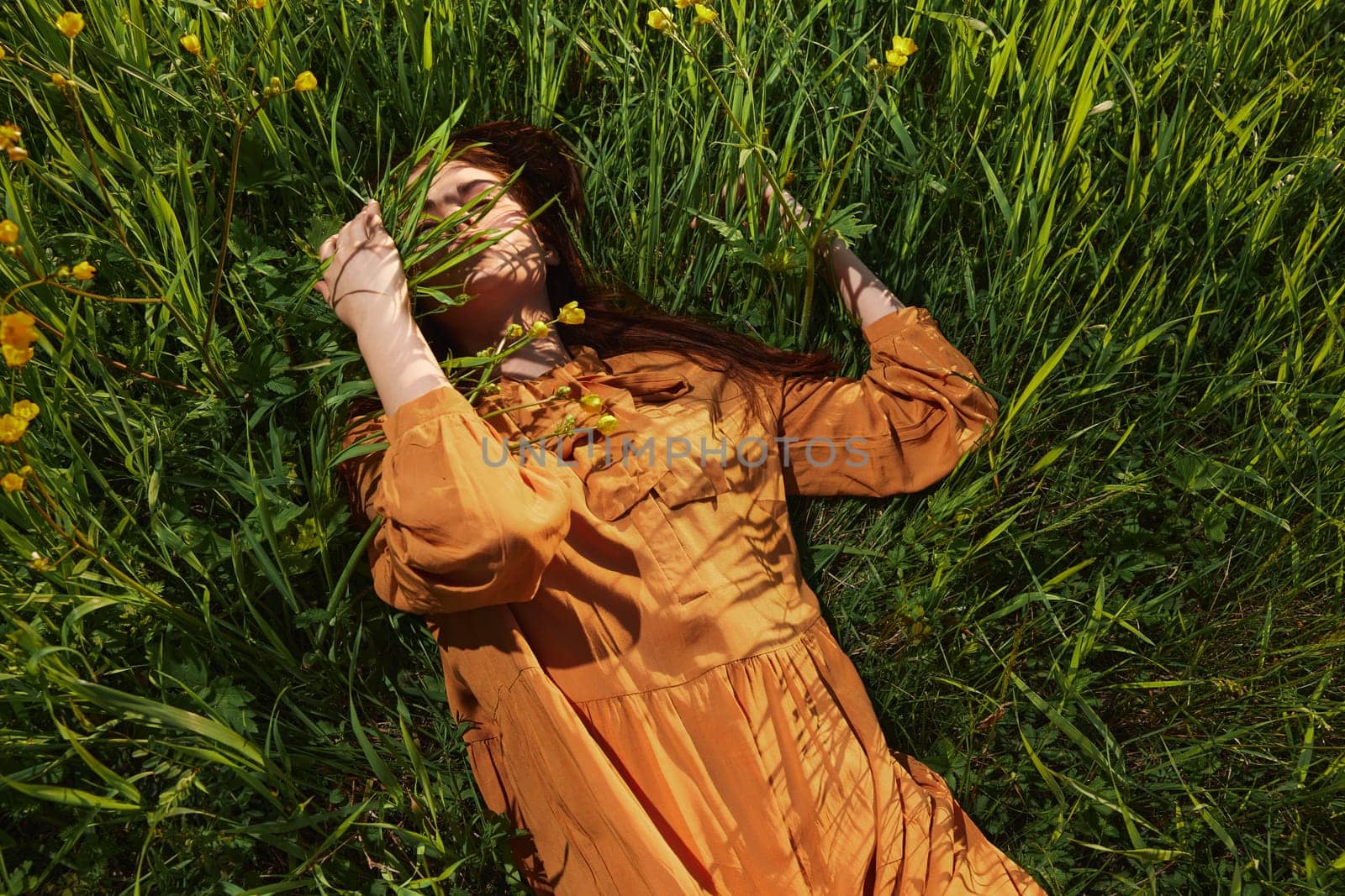 a calm woman with long red hair lies in a green field with yellow flowers, in an orange dress with her eyes closed, touching blades of grass with her hands, enjoying peace and recuperating. High quality photo