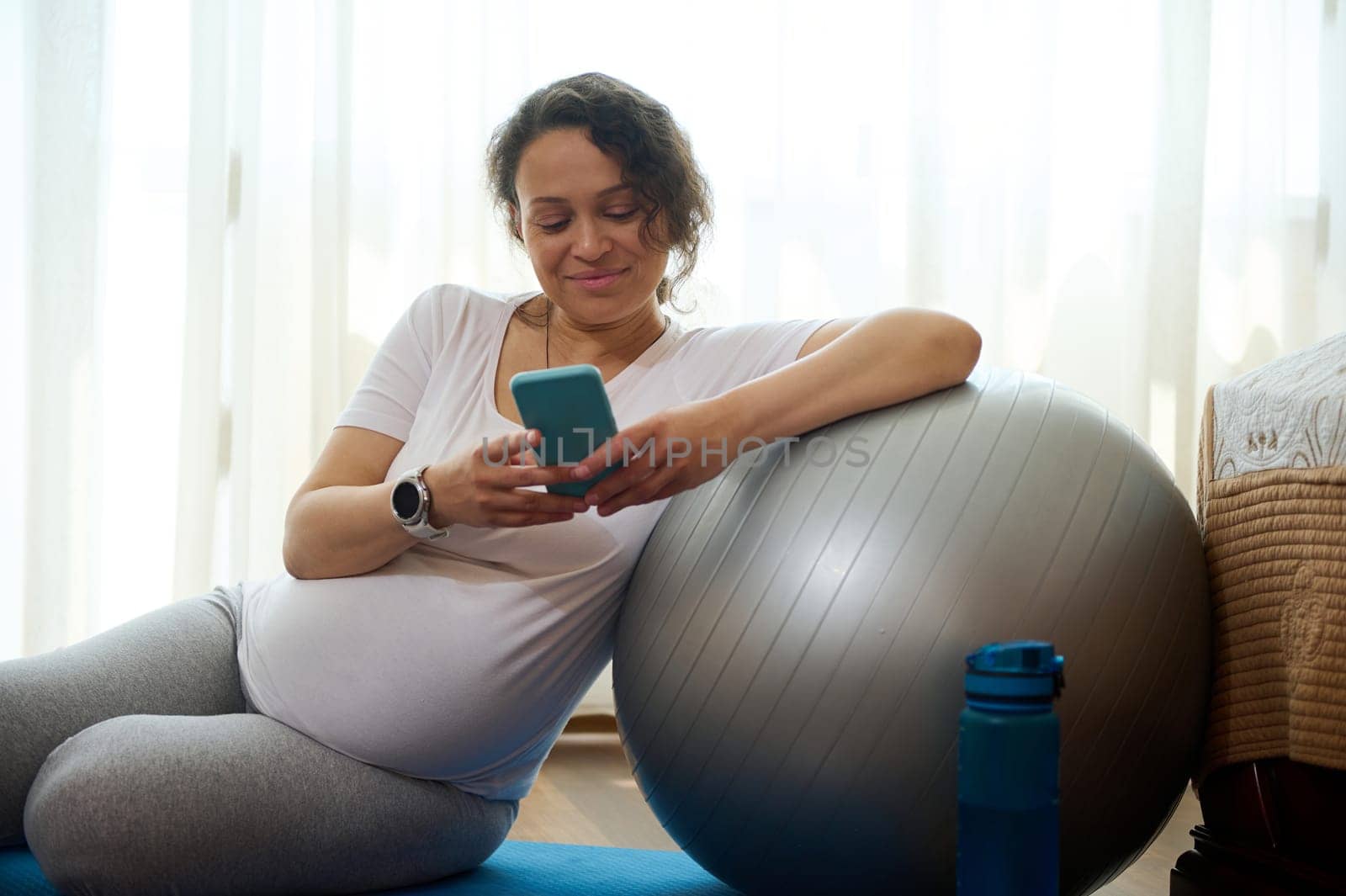 Happy young pregnant woman relaxes with smartphone on yoga mat after training with fit ball at home. Smiling expectant female browsing mobile application, resting after meditation practice, copy space