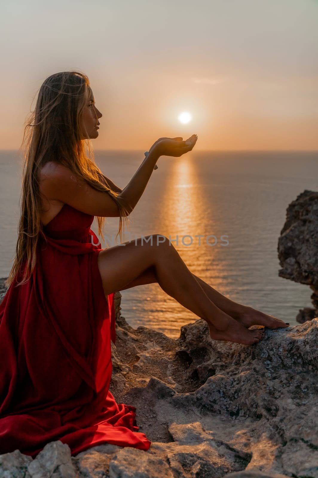 Woman sunset sea red dress, side view a happy beautiful sensual woman in a red long dress posing on a rock high above the sea on sunset