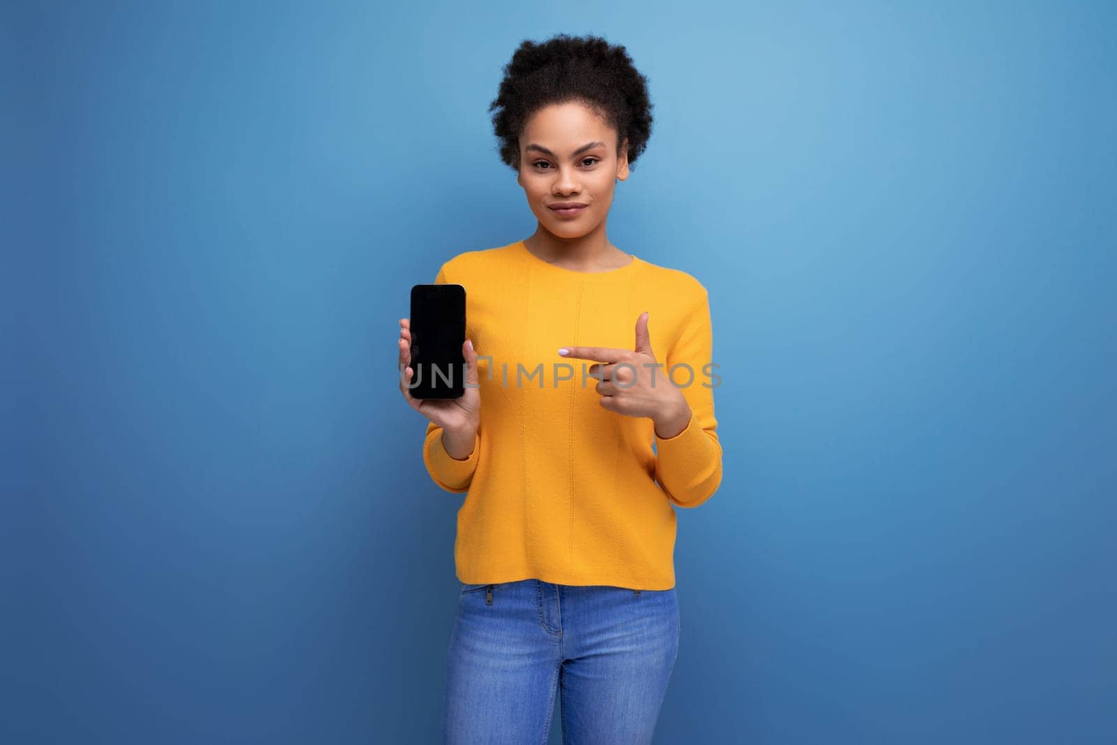 pretty 20s latin woman with afro hair in casual yellow blouse showing smartphone screen.