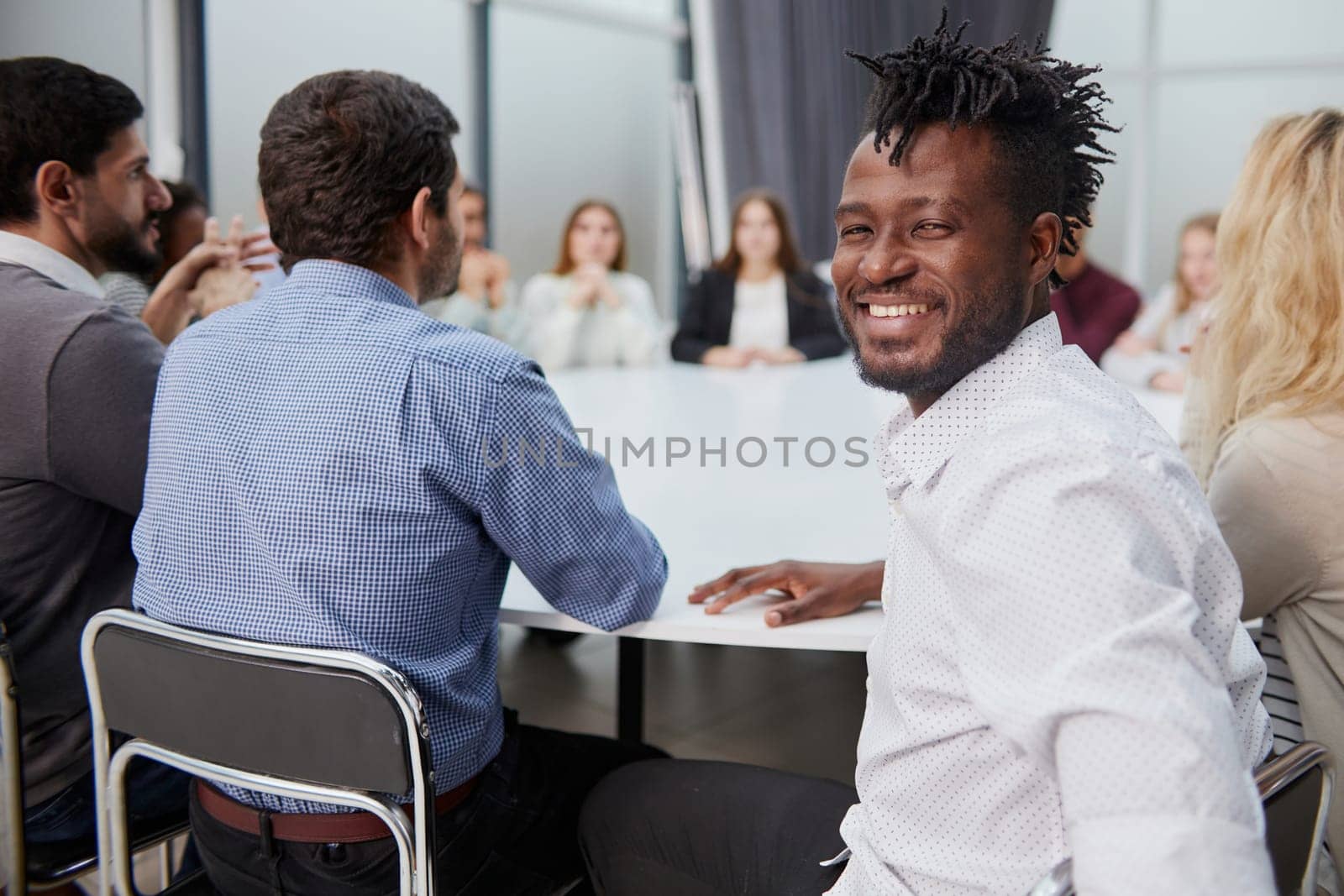 Portrait of a successful smiling young professional businessman sitting over desk in front of colleague working in background