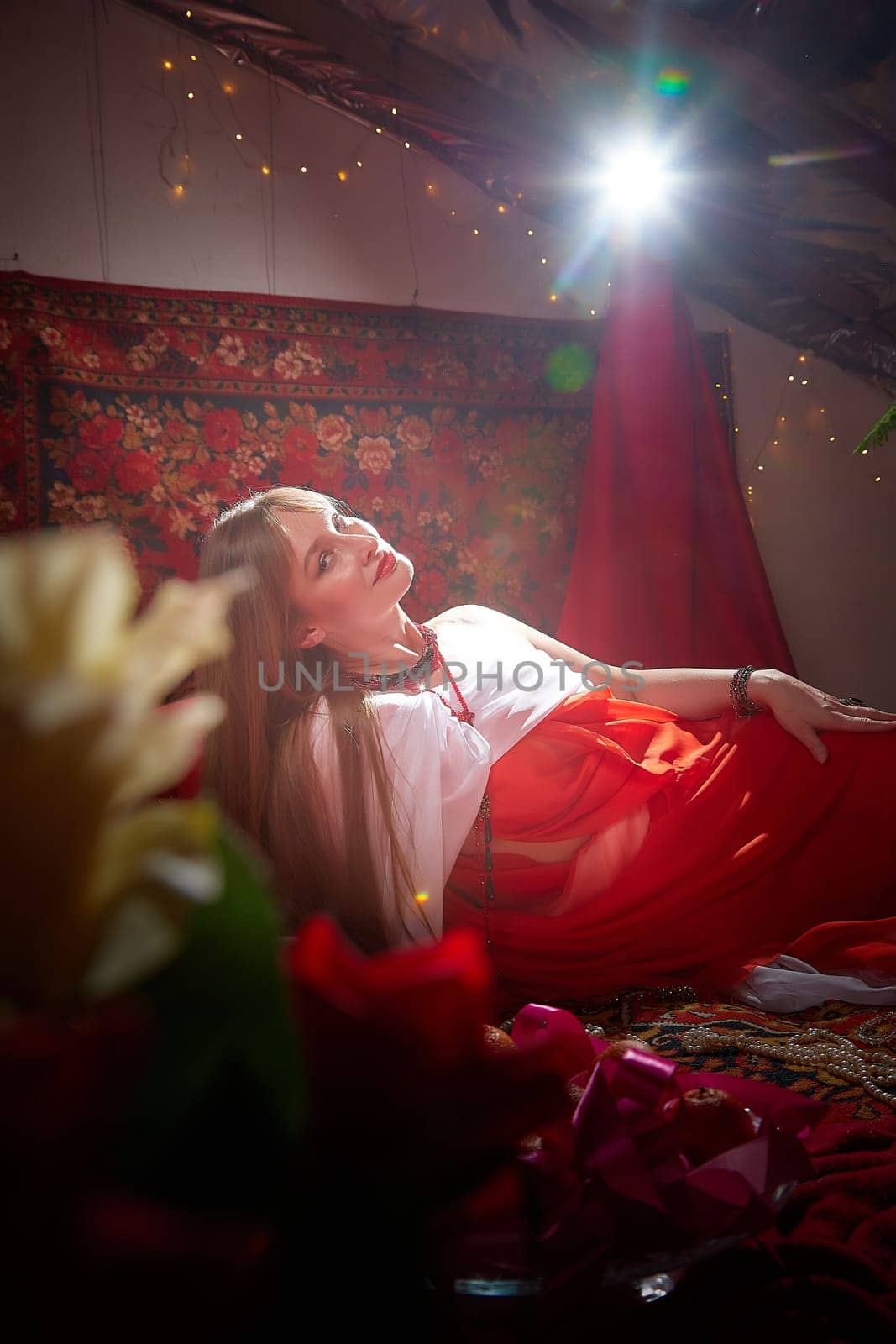 Beautiful European girl looking like Arab woman in red room with rich fabrics and carpets in sultan harem. Photo shoot of an oriental style odalisque. A model poses in sari as indian woman in india