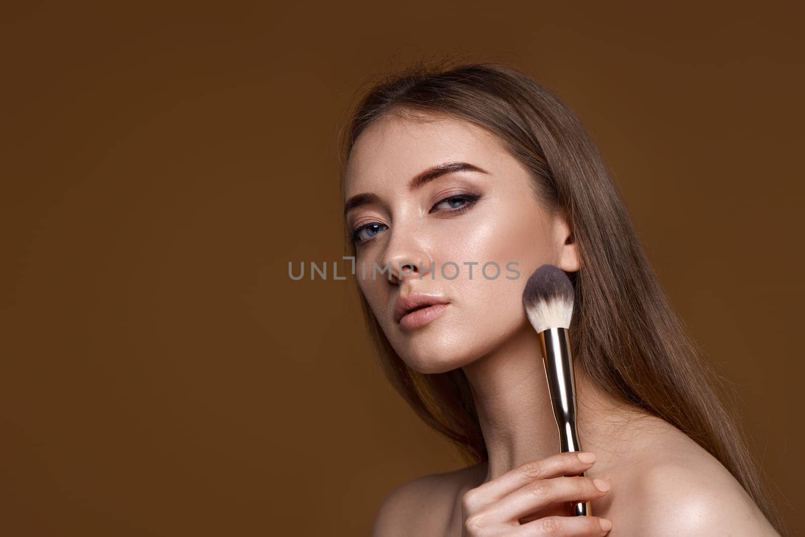 young woman applying blush on cheeks with make-up brush on beige background. copy space