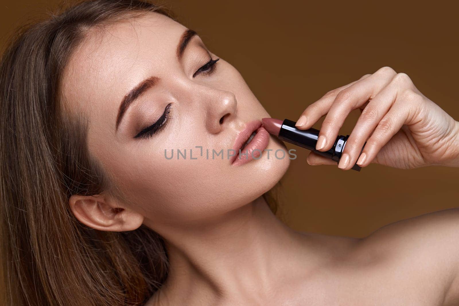 beautiful young smiling woman holding nude pink lipstick on beige background