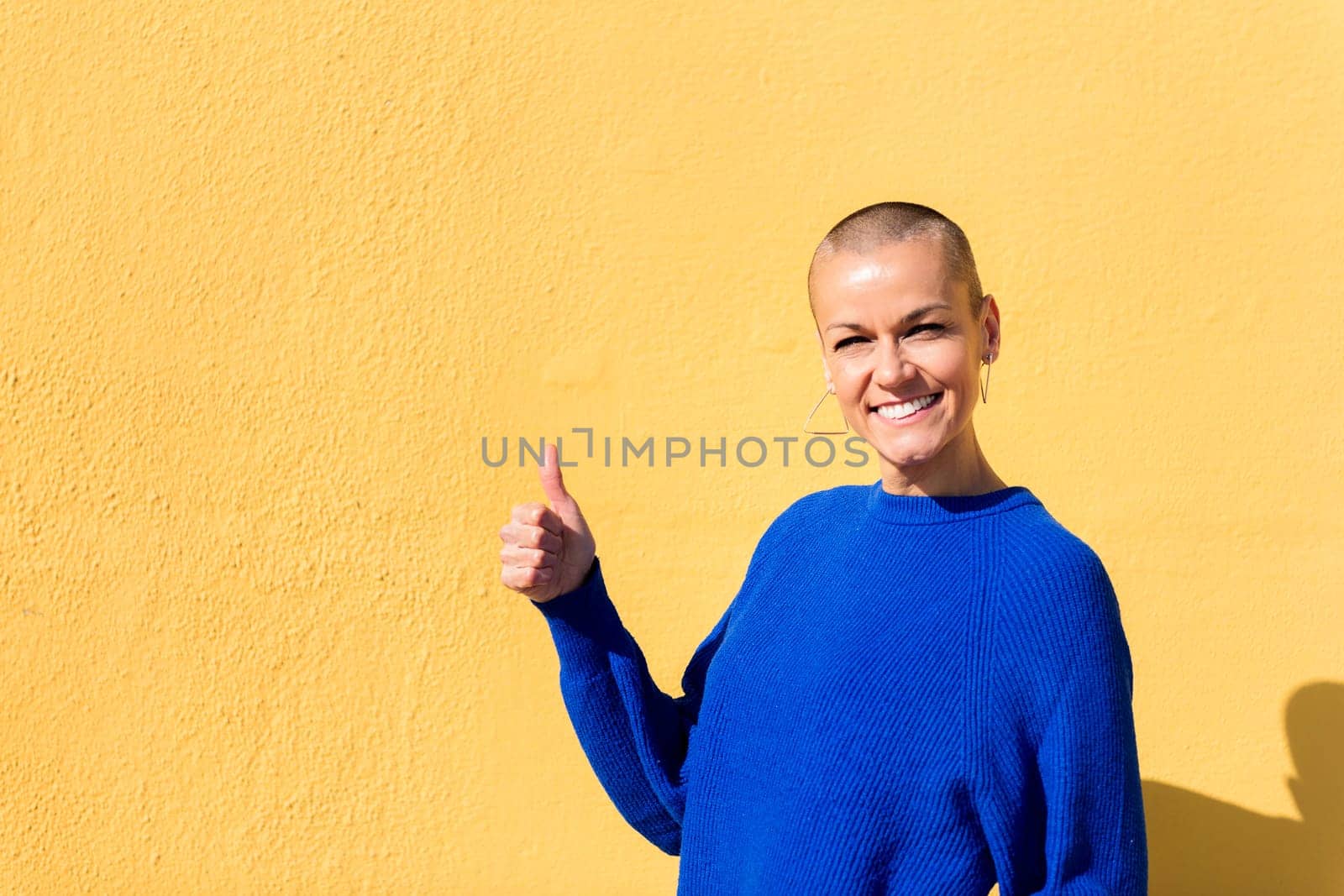 woman with a shaved head and wearing a blue sweater with her thumb up on a yellow wall in the background, concept of positive people, copy space for text