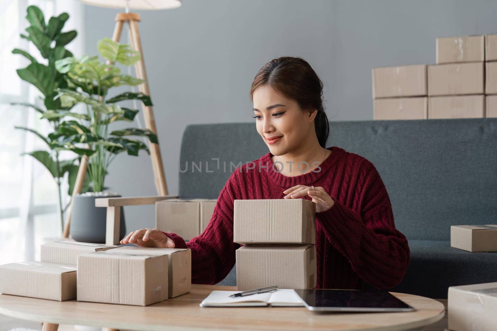 Asian woman holding parcel boxes and checking orders from tablet, she owns an online store, she packs and ships through a private transport company. Online selling and online shopping concepts.