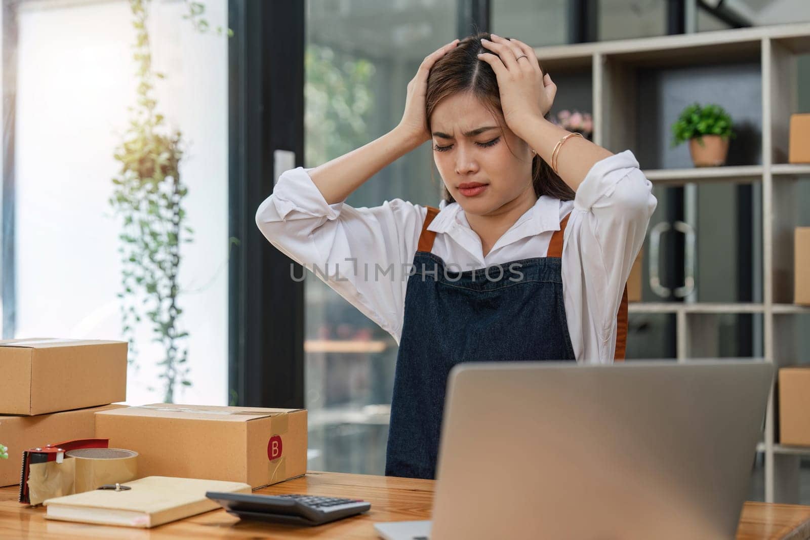 Small online business owner Asian woman wearing apron stressed headache taking notes with parcel box at home.