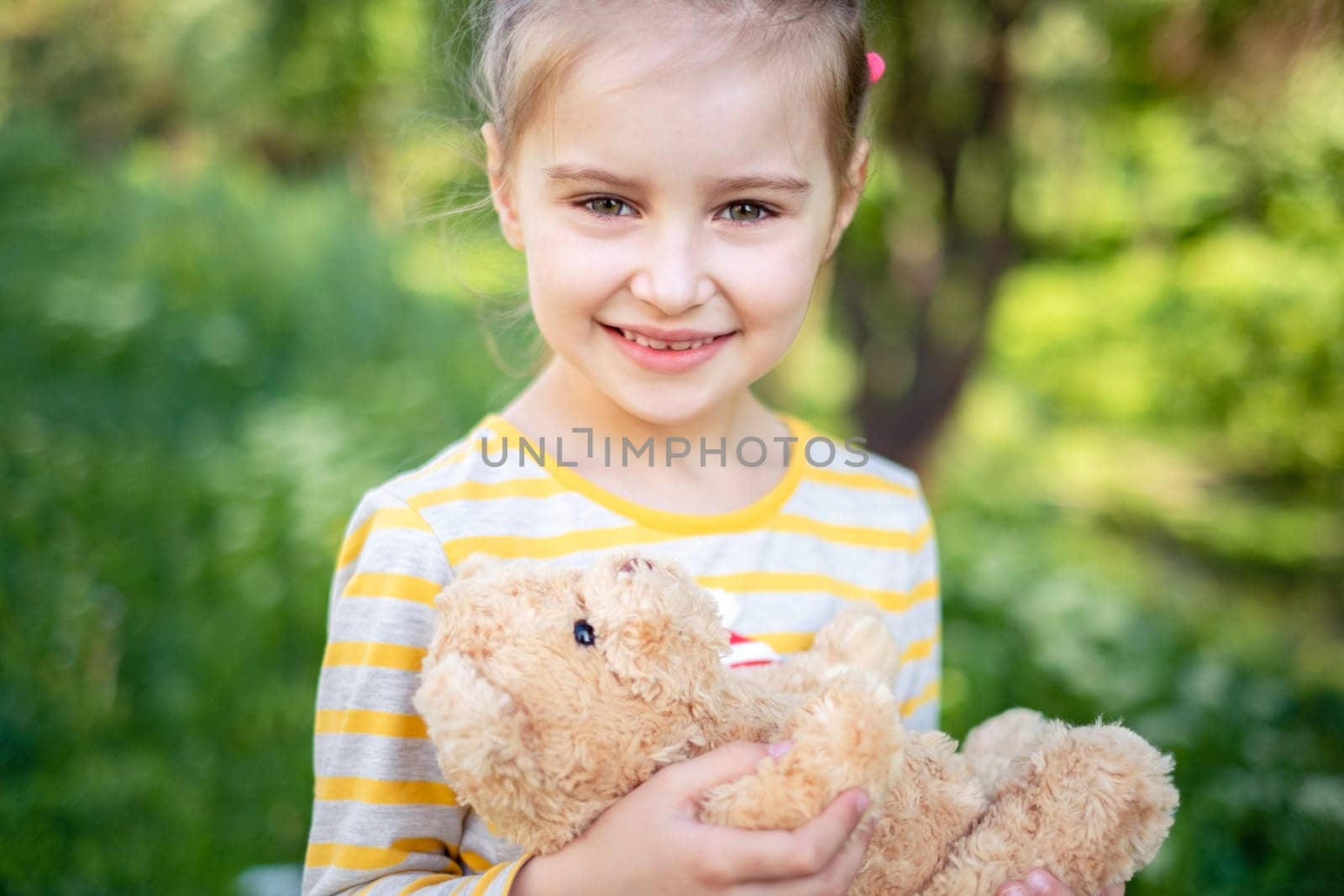 Cute smiling little child girl with her favorite teddy bear toy at the park in summer, looking ay camera