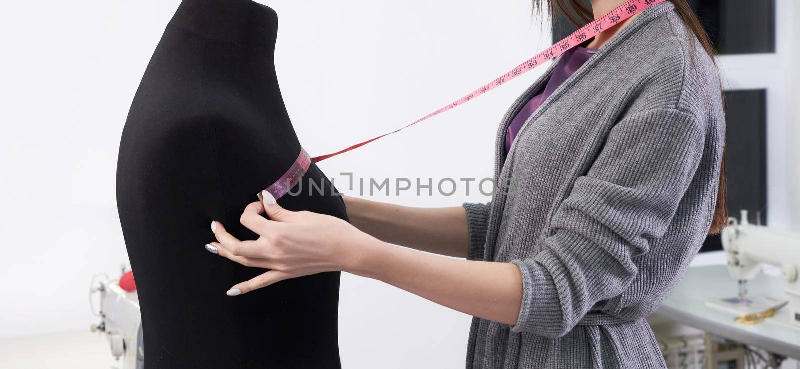Brunette seamstress in apron measuring beautiful fabric on black dummy in workshop
