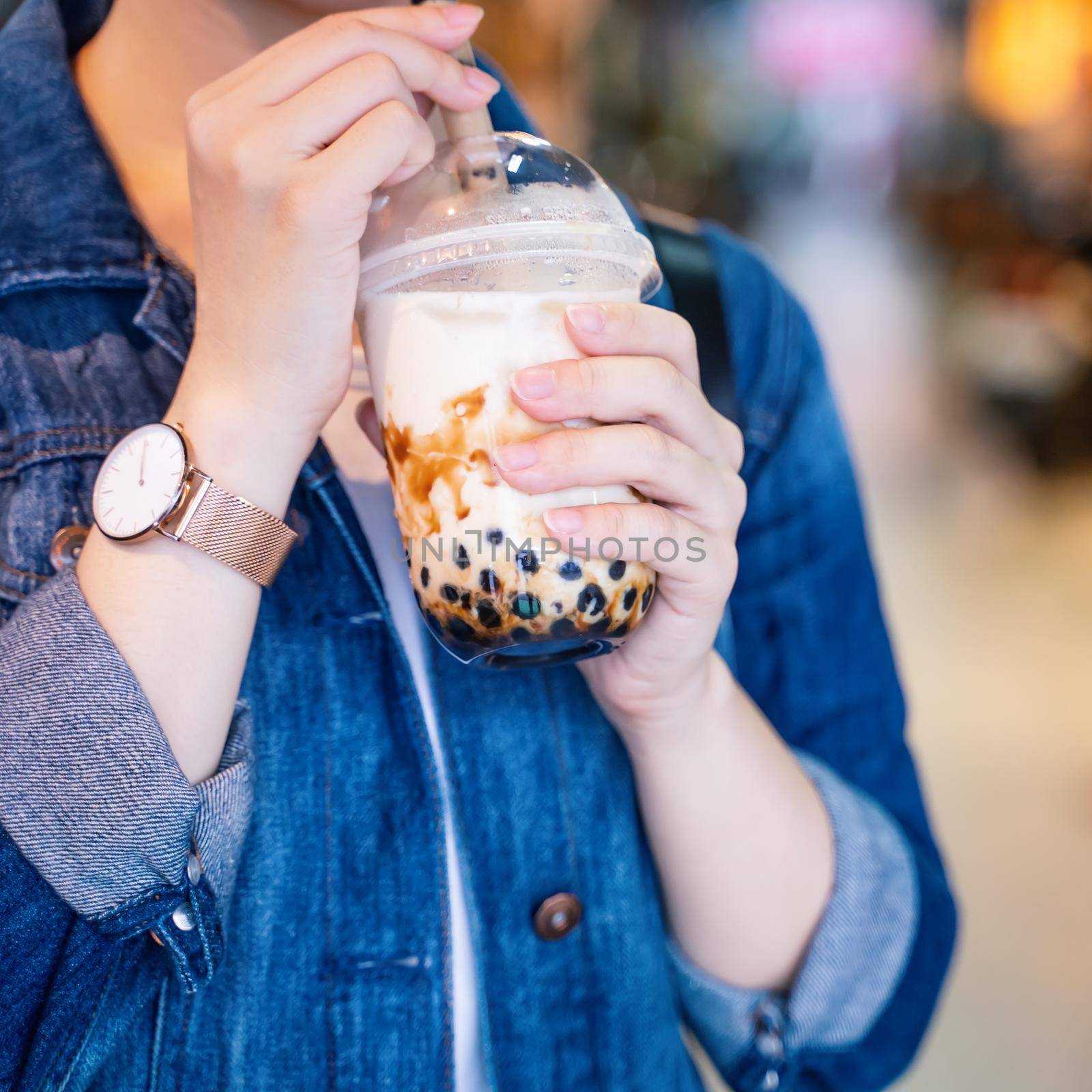 Young girl in denim jacket is drinking brown sugar flavored tapioca pearl bubble milk tea with glass straw in night market of Taiwan, close up, bokeh by ROMIXIMAGE
