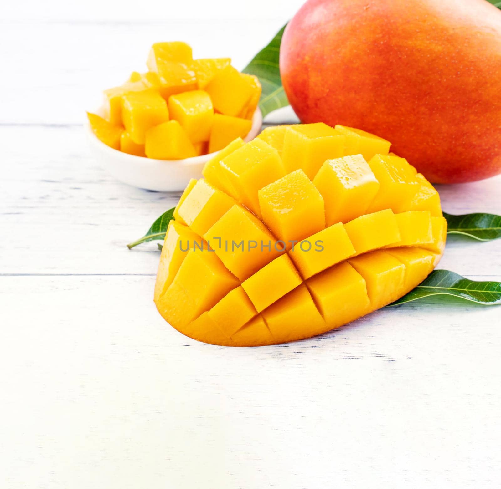 Beautiful chopped mango chunks with green leaves on bright white color timber background. Copy space, close up, macro. Tropical fruit concept.