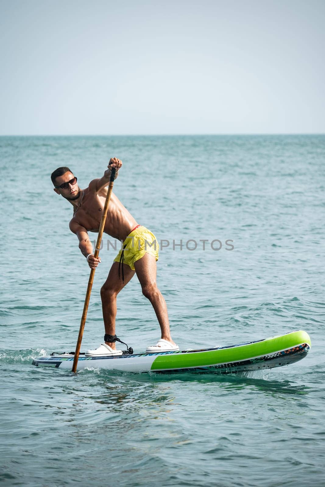 a sporty guy swims on a sup board with a paddle on the sea during the day against a beautiful sky by Rotozey