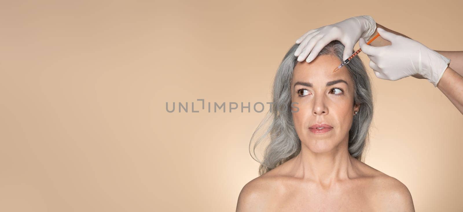 Senior caucasian woman on beauty procedure, cosmetologist hands in gloves making botox injection, beige background, panorama with free space. Anti-aging skin care, professional treatment