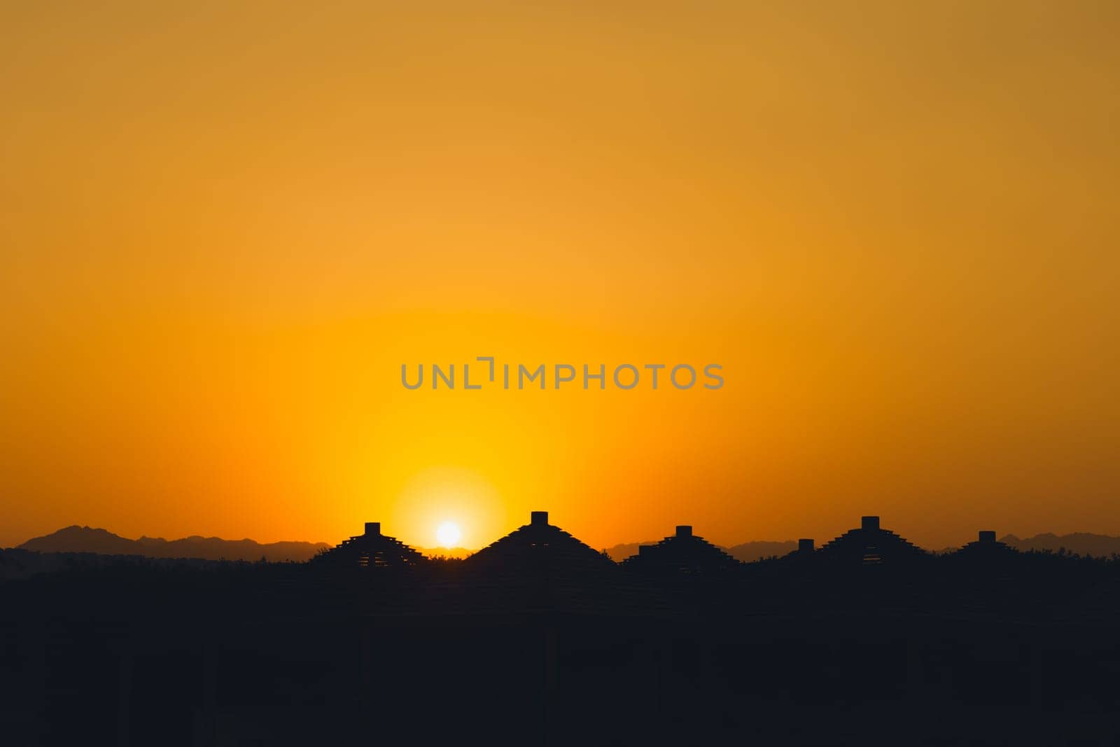 Majestic dusk in tropics. Goden sunset sky with beautiful silhouette roofs beach umbrellas and mountains in the evening. Warm orange colors. Abstract nature and travel background. Egypt summer