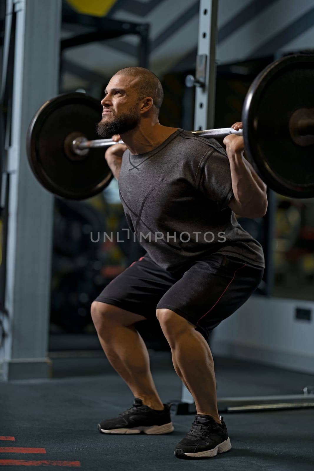 Strong muscular bearded short hair bodybuilder man holding a heavyweight barbell on the shoulder behind the neck while crouching and doing squats in the gym.