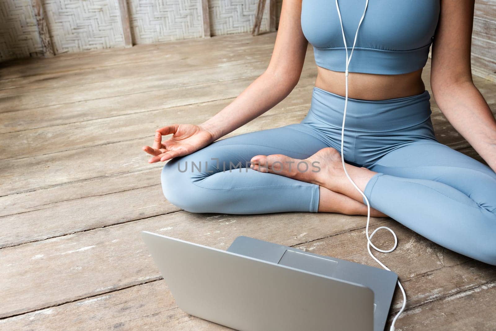 Unrecognisable woman wearing sports clothing doing online meditation using laptop and headphone sitting on wooden floor. Copy space. Spirituality, mental health and technology concepts.