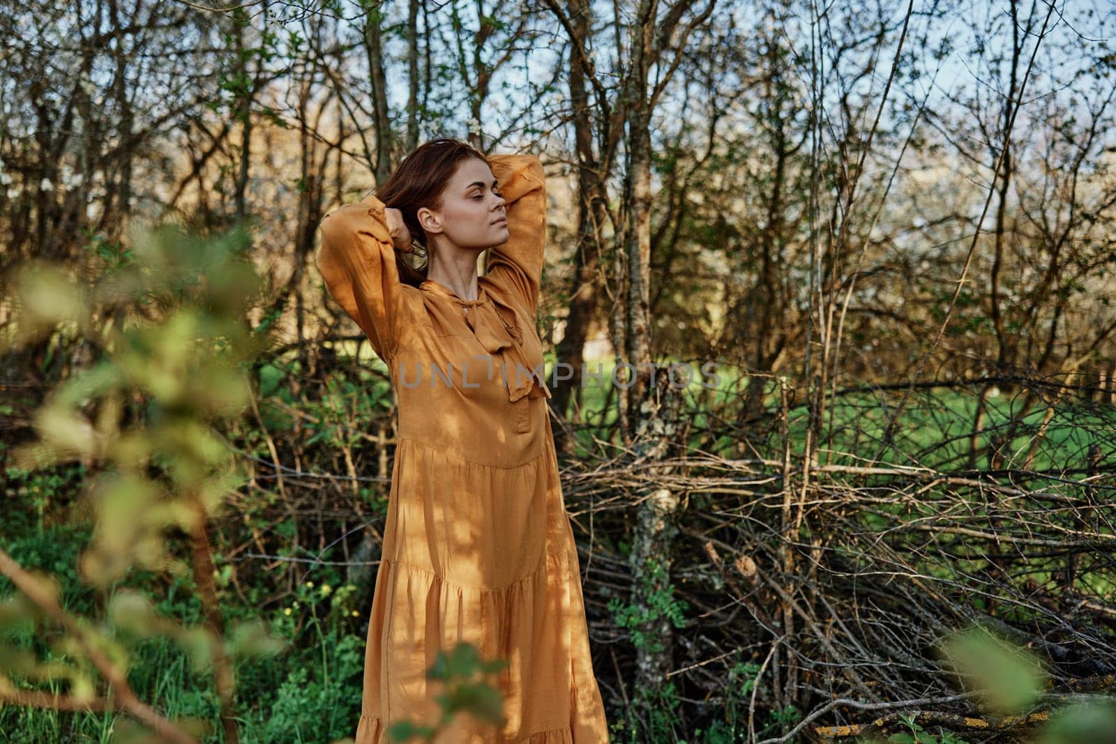 a beautiful, slender woman with long hair walks in the shade near the trees, dressed in a long orange dress, enjoying the weather and the weekend, straightening her hair with her hands by Vichizh