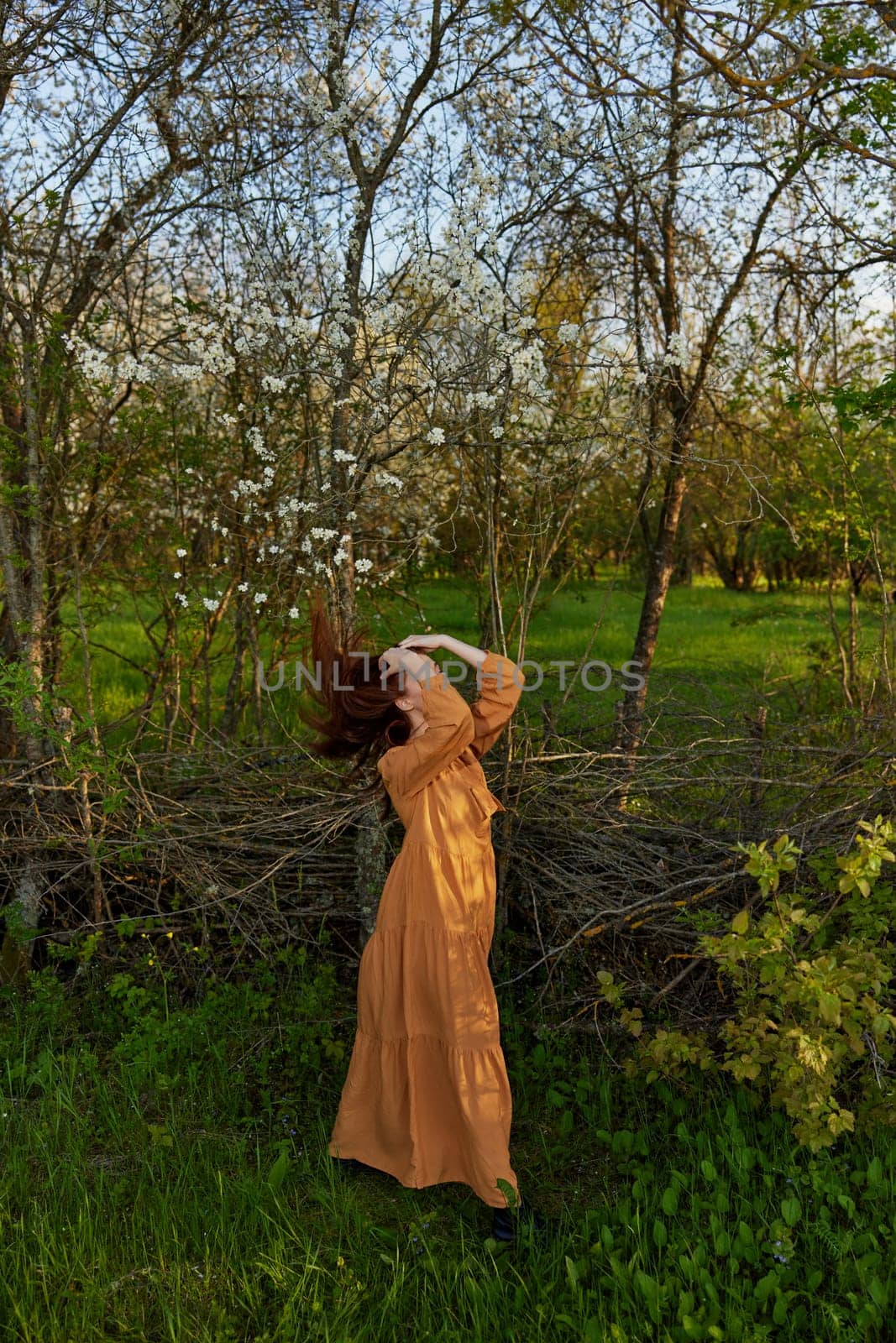 an elegant, sophisticated woman poses relaxed standing near a wicker fence at the dacha in a long orange dress tossing her long red hair. High quality photo