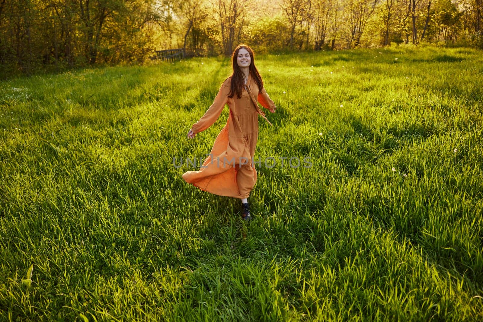 slender, red-haired woman stands in a wide, green field during sunset in a long orange dress enjoying unity with nature and relaxation, raising her arms to the sides while facing the camera by Vichizh