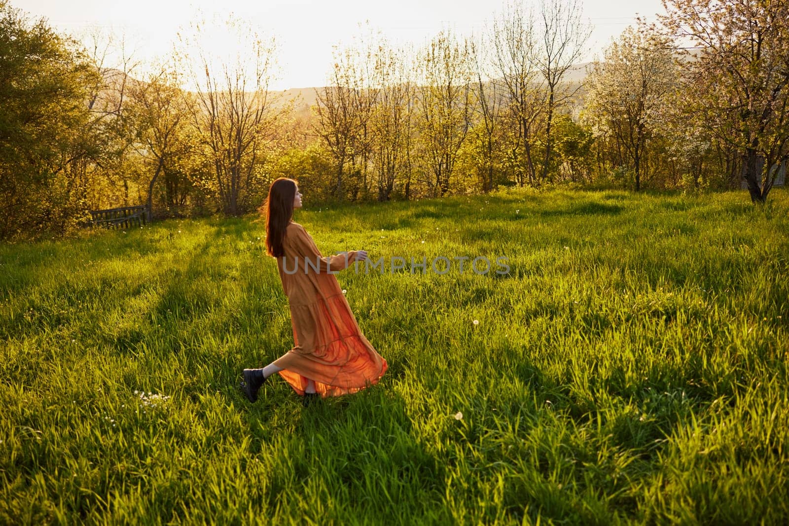 an attractive, slender, red-haired woman stands in a wide green field during sunset in a long orange dress enjoying unity with nature and relaxation by Vichizh